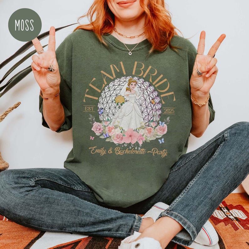 May include: A green t-shirt with the text "Team Bride Est 2023" and a graphic of a princess in a white dress standing in front of a purple and gold sphere. The text "Emily & Bachelorette Party" is below the graphic.