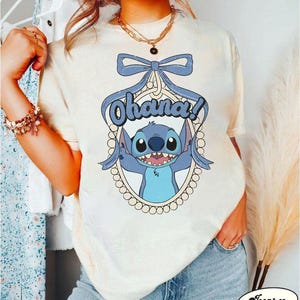 Puede incluir: Una camiseta blanca con un personaje azul de Stitch de Lilo & Stitch. Stitch sostiene un cartel que dice "Ohana!" y est&aacute; rodeado de un marco de perlas. La camiseta tiene un aspecto desgastado y un lazo en la parte superior.