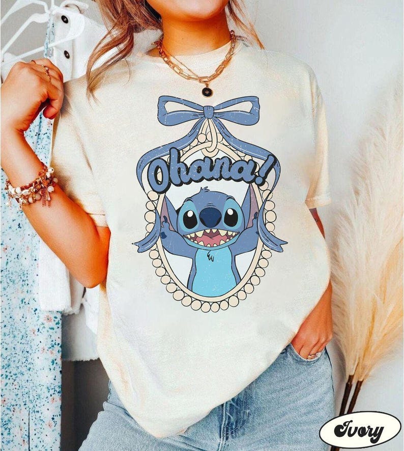 Puede incluir: Una camiseta blanca con un personaje azul de Stitch de Lilo & Stitch. Stitch sostiene un cartel que dice "Ohana!" y est&aacute; rodeado de un marco de perlas. La camiseta tiene un aspecto desgastado y un lazo en la parte superior.