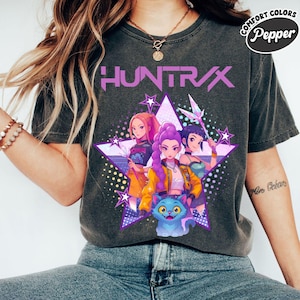 Pode incluir: T-shirt cinza escuro com a palavra "HUNTRAX" em roxo, com um gr&aacute;fico de tr&ecirc;s mulheres em estilo anime dentro de uma estrela roxa. As mulheres est&atilde;o em v&aacute;rias roupas, e um gato azul est&aacute; na parte inferior.