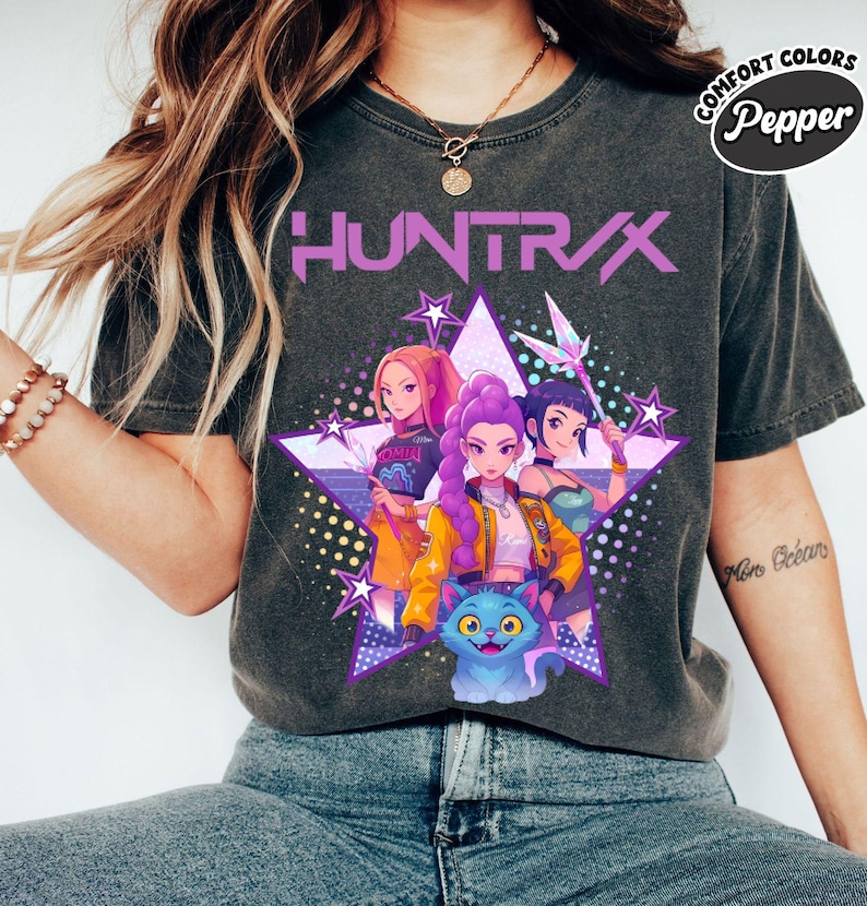 Pode incluir: T-shirt cinza escuro com a palavra "HUNTRAX" em roxo, com um gr&aacute;fico de tr&ecirc;s mulheres em estilo anime dentro de uma estrela roxa. As mulheres est&atilde;o em v&aacute;rias roupas, e um gato azul est&aacute; na parte inferior.