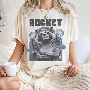 Puede incluir: Una camiseta de color crema con un gráfico de Rocket Raccoon de Guardianes de la Galaxia. El gráfico es una ilustración de estilo vintage de Rocket en su traje espacial, con la palabra "ROCKET" impresa encima.