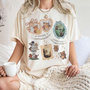 Puede incluir: Una camiseta de color crema con un estampado gráfico de varios personajes de Star Wars. Los personajes están rodeados de pequeñas cadenas doradas con cuentas y las palabras "A Tribe Called Quest" y "The Most Beautiful" impresas en ellas.