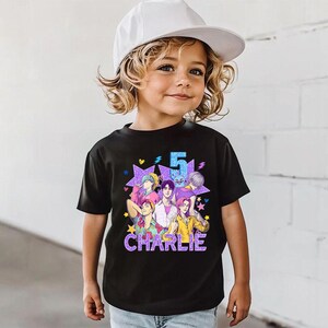 May include: Black graphic t-shirt featuring a colorful anime-style design with the number 5 and the name "Charlie". The design includes multiple figures and stars. The child is wearing a white baseball cap.