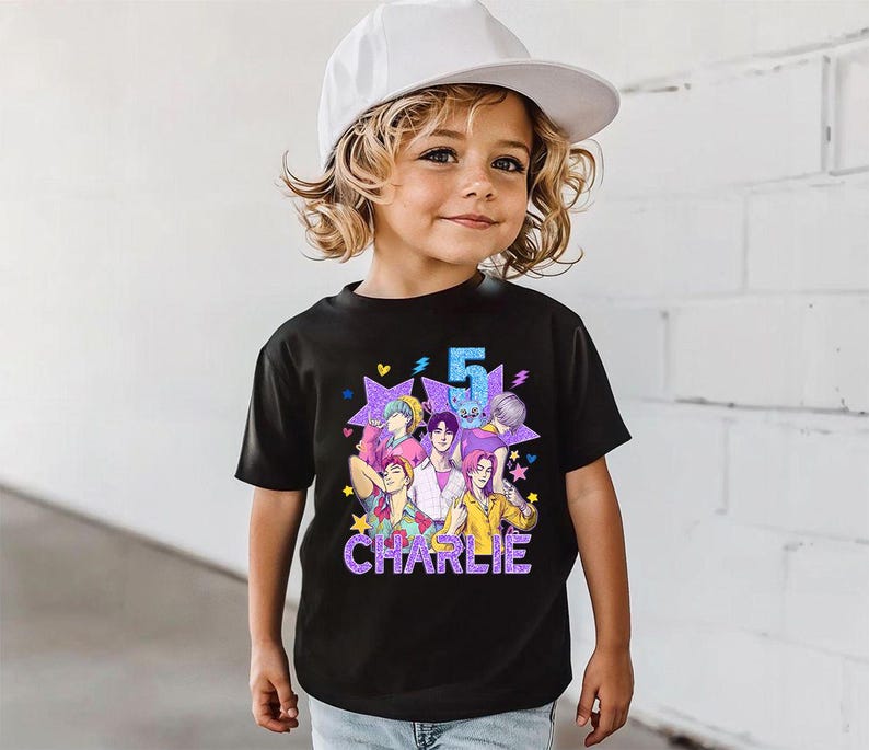 May include: Black graphic t-shirt featuring a colorful anime-style design with the number 5 and the name "Charlie". The design includes multiple figures and stars. The child is wearing a white baseball cap.