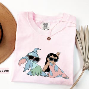 May include: Pink t-shirt with a graphic print of Stitch and Lilo from the Disney movie Lilo & Stitch. Stitch is wearing sunglasses and a green grass skirt. Lilo is wearing sunglasses and a pink dress. The shirt has a Comfort Colors tag.
