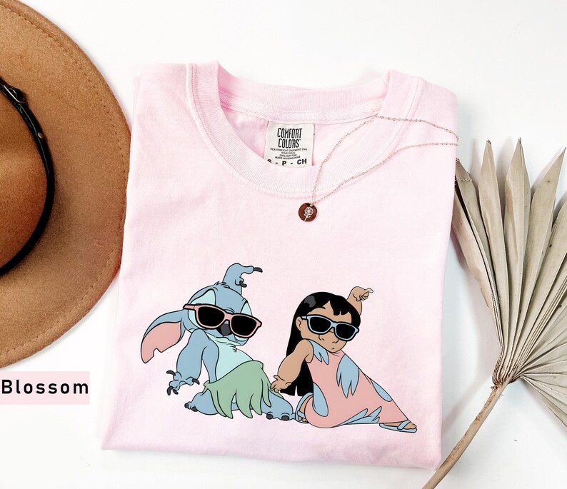 May include: Pink t-shirt with a graphic print of Stitch and Lilo from the Disney movie Lilo & Stitch. Stitch is wearing sunglasses and a green grass skirt. Lilo is wearing sunglasses and a pink dress. The shirt has a Comfort Colors tag.
