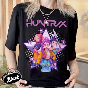 Pode incluir: Camiseta preta com um gr&aacute;fico de tr&ecirc;s mulheres em estilo anime e um gato azul, em um design de estrela roxa. A palavra "HUNTR/X" &eacute; impressa acima do gr&aacute;fico em roxo. A camisa tem as palavras "COMFORT COLORS" e "Black".