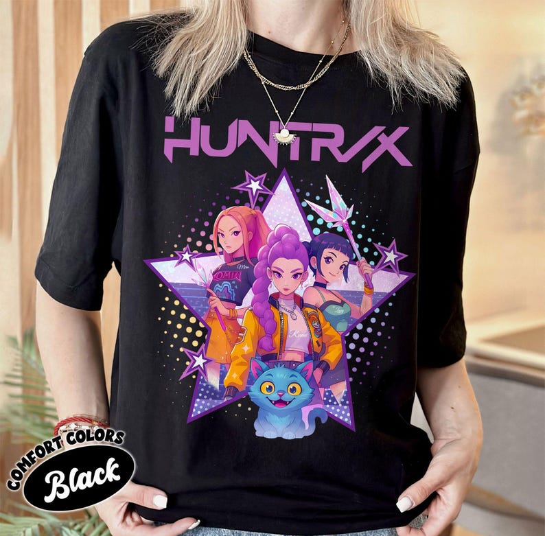 Pode incluir: Camiseta preta com um gr&aacute;fico de tr&ecirc;s mulheres em estilo anime e um gato azul, em um design de estrela roxa. A palavra "HUNTR/X" &eacute; impressa acima do gr&aacute;fico em roxo. A camisa tem as palavras "COMFORT COLORS" e "Black".