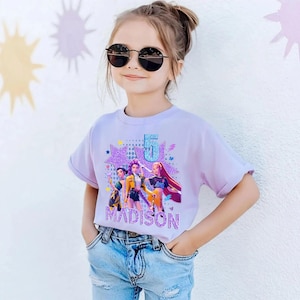 May include: A young girl wearing a light purple t-shirt with a graphic design. The design features a number 5, three cartoon figures, and the name "MADISON" in a glittery font. She is also wearing sunglasses and blue jeans.