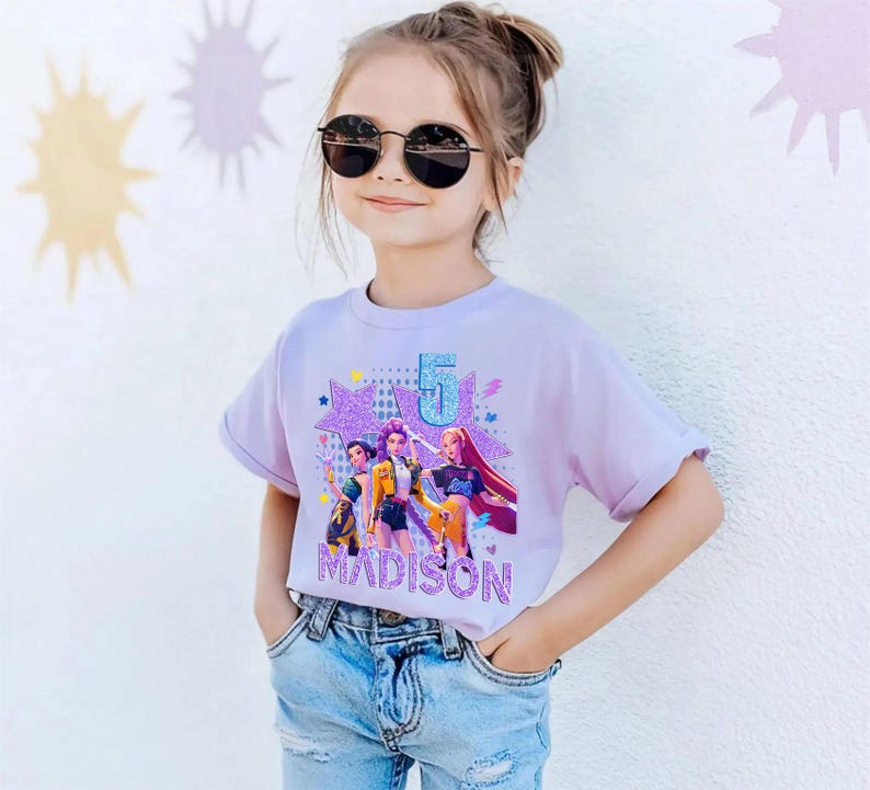 May include: A young girl wearing a light purple t-shirt with a graphic design. The design features a number 5, three cartoon figures, and the name "MADISON" in a glittery font. She is also wearing sunglasses and blue jeans.