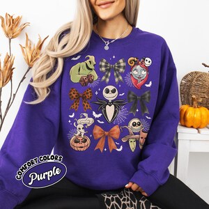 May include: A purple Comfort Colors sweatshirt with a design featuring characters and bows from The Nightmare Before Christmas. The design includes Jack Skellington, Sally, Oogie Boogie, and other Halloween-themed elements. The text "Comfort Colors Purple" is printed on the lower left.