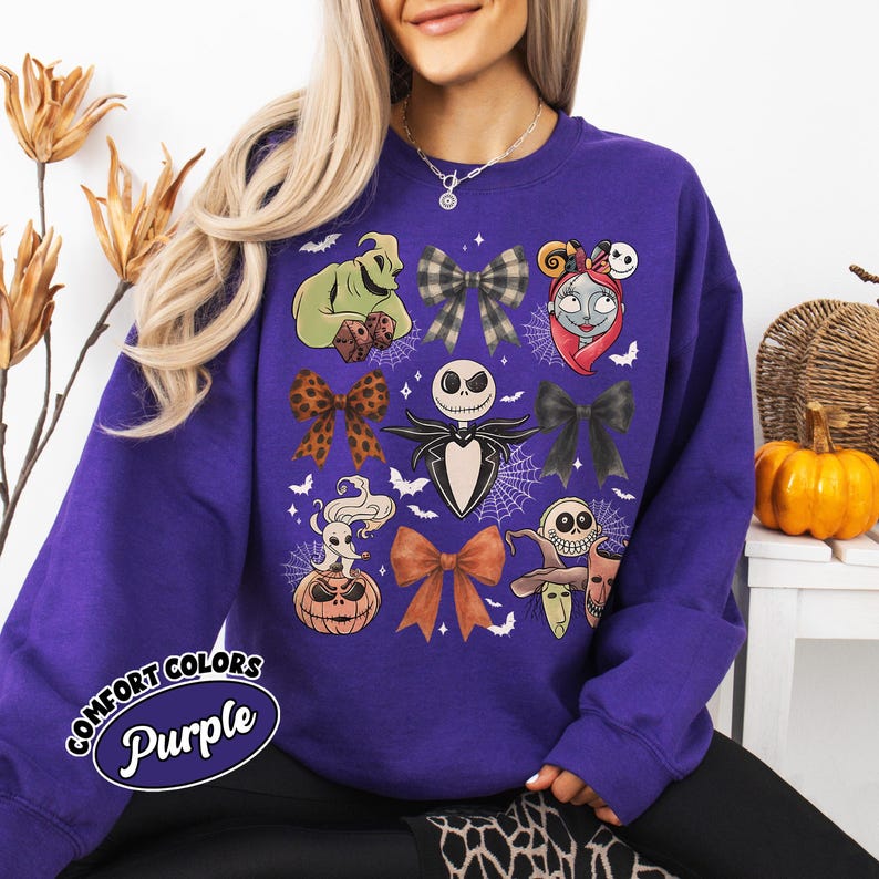 May include: A purple Comfort Colors sweatshirt with a design featuring characters and bows from The Nightmare Before Christmas. The design includes Jack Skellington, Sally, Oogie Boogie, and other Halloween-themed elements. The text "Comfort Colors Purple" is printed on the lower left.