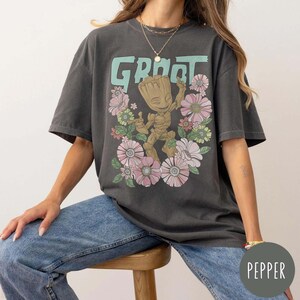May include: A dark grey t-shirt featuring a cartoon character called Groot from the Guardians of the Galaxy film series. The character is surrounded by pink flowers and the word "Groot" is printed in green above the character. The shirt is being worn by a person wearing blue jeans.