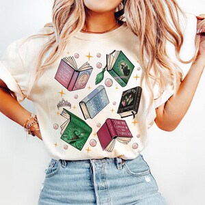 May include: Cream-colored t-shirt featuring a whimsical design of open books with various titles, including "Defying Gravity" and "You're Gonna Be Popular." The design includes pink bubbles, stars, and a tiara.