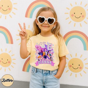 May include: A young girl wearing heart-shaped sunglasses and a yellow t-shirt with a colorful graphic design. The shirt features the number 6, the name "GIANNA", and images of animated characters. The background includes rainbows and smiling suns.