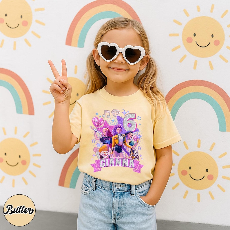 May include: A young girl wearing heart-shaped sunglasses and a yellow t-shirt with a colorful graphic design. The shirt features the number 6, the name "GIANNA", and images of animated characters. The background includes rainbows and smiling suns.