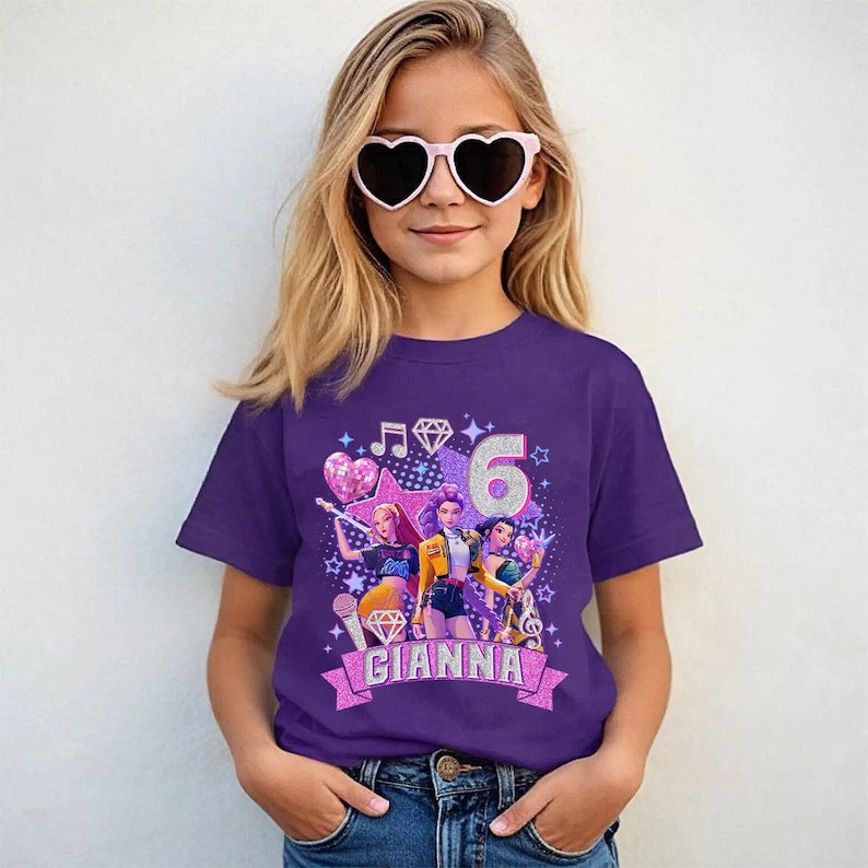 May include: A purple t-shirt featuring a graphic design with the number 6, musical notes, stars, and three cartoon characters. The name "Gianna" is displayed on a banner. The girl is wearing heart-shaped sunglasses.