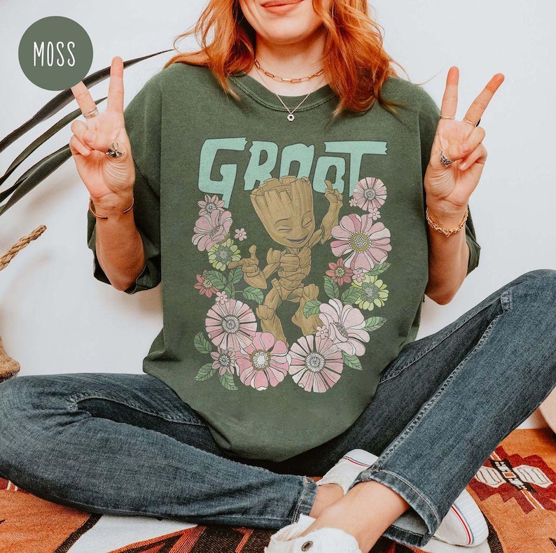 May include: A woman wearing a green t-shirt featuring a graphic of Groot from Guardians of the Galaxy dancing amongst flowers. The shirt reads "Groot" in large letters.
