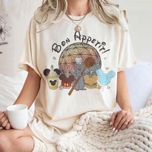 Op de afbeelding: Een crèmekleurig T-shirt met een grafische print van een kokrat, Mickey Mouse en een eetkraam. De tekst "Bon Appetit!" staat op het shirt gedrukt.