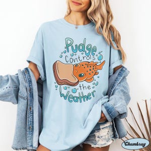 May include: Light blue t-shirt featuring a cartoon illustration of a fish eating a slice of bread. The text on the shirt reads "Pudge controls the weather".