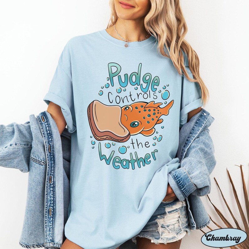 May include: Light blue t-shirt featuring a cartoon illustration of a fish eating a slice of bread. The text on the shirt reads "Pudge controls the weather".
