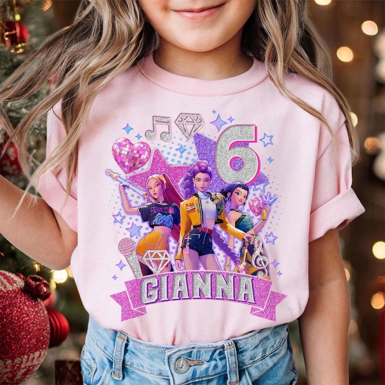 May include: Pink t-shirt with a graphic design featuring three animated figures, musical notes, stars, and the number 6. The name "GIANNA" is displayed on a banner. The design includes glitter accents.