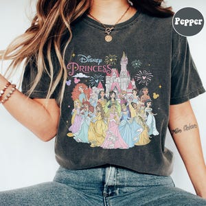 May include: A dark gray t-shirt with a vintage look featuring a colorful illustration of Disney princesses in front of Cinderella's castle. The text "Disney Princess" is printed in pink and white above the illustration.