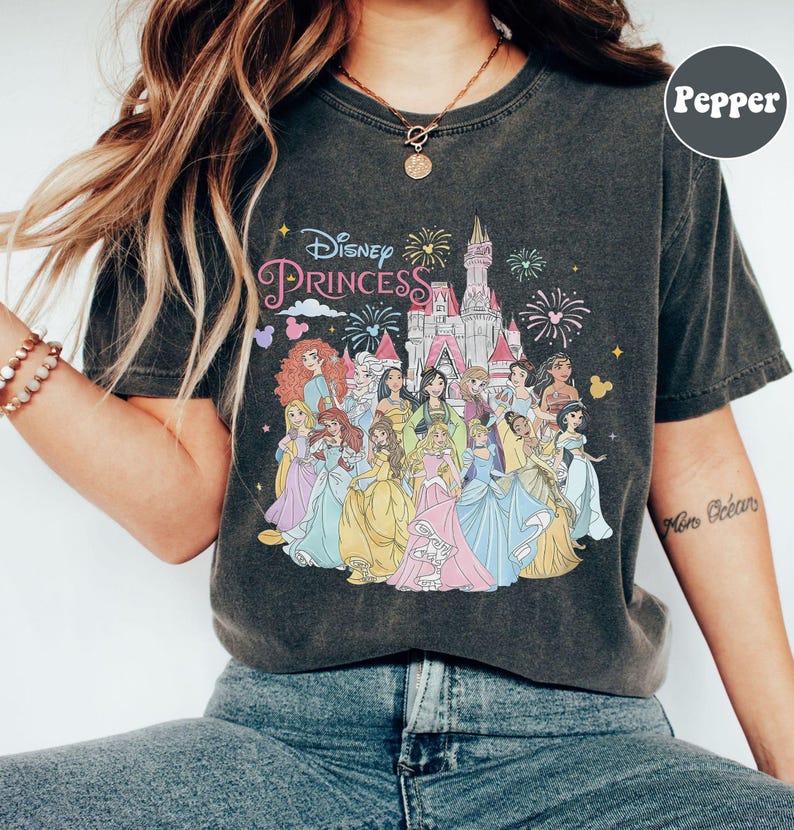 May include: A dark gray t-shirt with a vintage look featuring a colorful illustration of Disney princesses in front of Cinderella's castle. The text "Disney Princess" is printed in pink and white above the illustration.