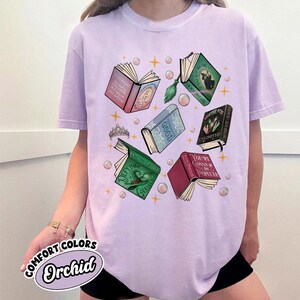 May include: Orchid-colored t-shirt featuring a graphic of open books with titles like "Defying Gravity" and "You're Gonna Be Popular." The design includes a tiara, bubbles, and stars. The shirt is labeled "Comfort Colors."