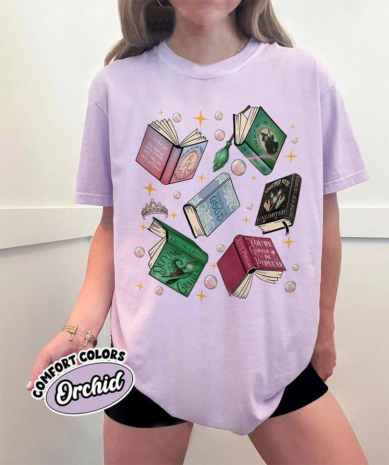 May include: Orchid-colored t-shirt featuring a graphic of open books with titles like "Defying Gravity" and "You're Gonna Be Popular." The design includes a tiara, bubbles, and stars. The shirt is labeled "Comfort Colors."