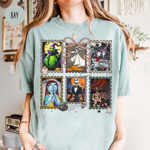 May include: A light blue t-shirt featuring a collage of six postage stamp-style images of characters from The Nightmare Before Christmas. The stamps include images of Jack Skellington, Sally, and Zero. The word "BAY" is printed on the upper left.