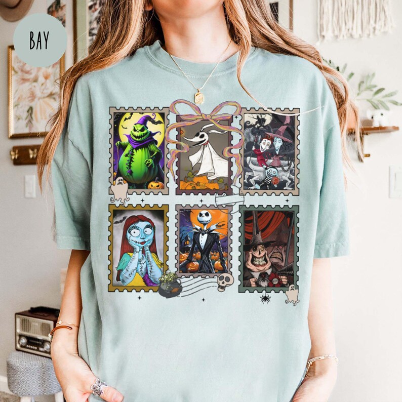 May include: A light blue t-shirt featuring a collage of six postage stamp-style images of characters from The Nightmare Before Christmas. The stamps include images of Jack Skellington, Sally, and Zero. The word "BAY" is printed on the upper left.