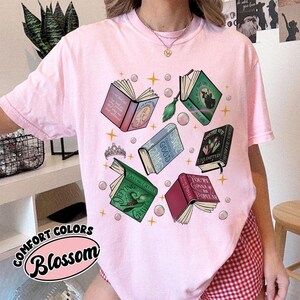 May include: Pink t-shirt with a graphic of open books, a tiara, and bubbles. The books have titles like "Defying Gravity" and "You're Gonna Be Popular." The text "COMFORT COLORS Blossom" is also visible.