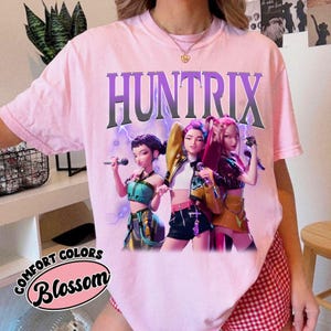Pu&ograve; includere: T-shirt rosa chiaro con la scritta "HUNTRIX" in lettere viola. La maglietta presenta una grafica di tre figure animate che tengono dei microfoni. Il testo "COMFORT COLORS Blossom" &egrave; stampato in basso a sinistra.