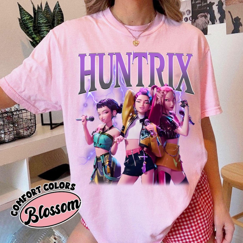 Pu&ograve; includere: T-shirt rosa chiaro con la scritta "HUNTRIX" in lettere viola. La maglietta presenta una grafica di tre figure animate che tengono dei microfoni. Il testo "COMFORT COLORS Blossom" &egrave; stampato in basso a sinistra.