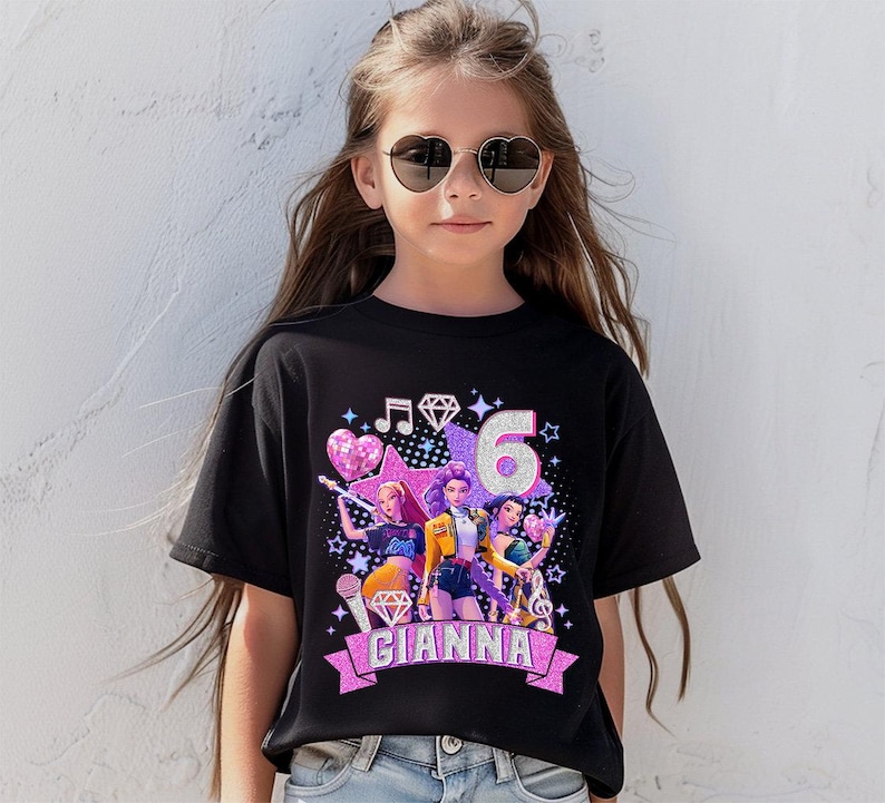 May include: Black t-shirt with a colorful graphic featuring three animated figures, musical notes, and the number 6. The name "GIANNA" is displayed on a pink banner. The design includes stars, hearts, and diamonds.