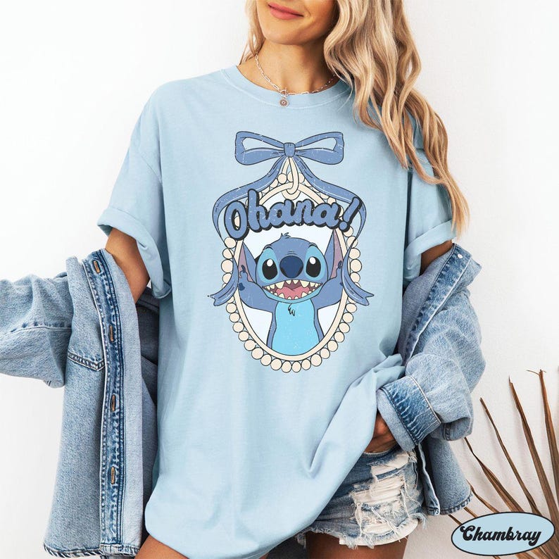 Puede incluir: Una camiseta azul claro con un personaje azul Stitch sosteniendo un cartel que dice "Ohana!" Stitch est&aacute; rodeado de un collar de perlas y un lazo azul.
