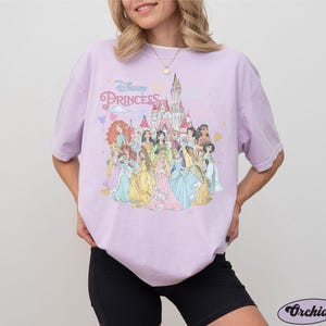 May include: A lavender t-shirt with a graphic print of Disney princesses in front of Cinderella's castle. The text "Disney Princess" is printed on the shirt.