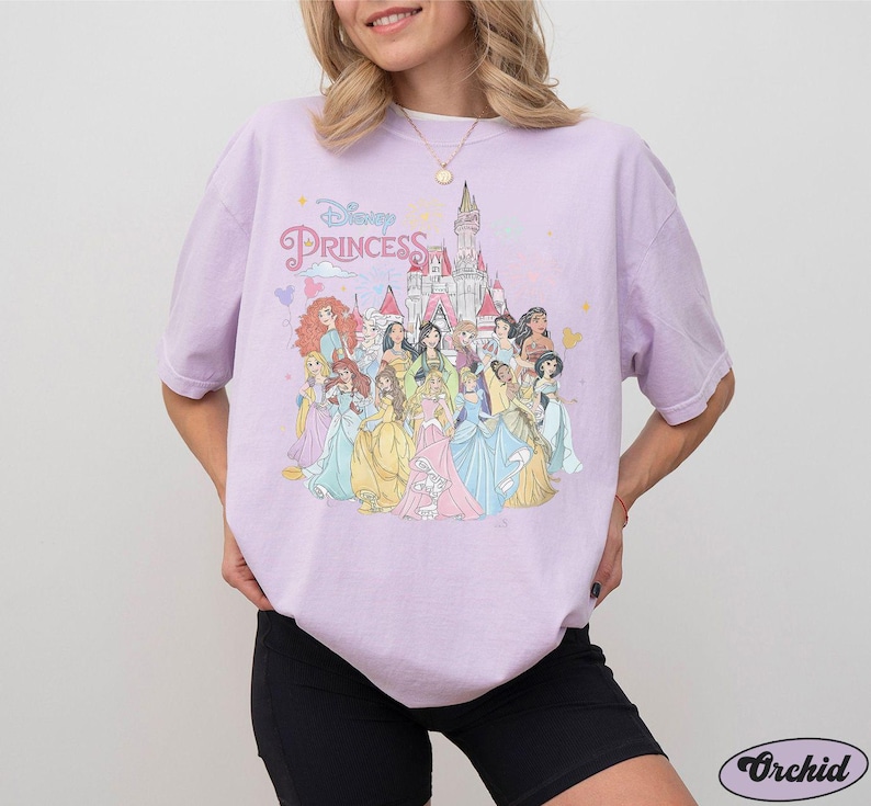 May include: A lavender t-shirt with a graphic print of Disney princesses in front of Cinderella's castle. The text "Disney Princess" is printed on the shirt.