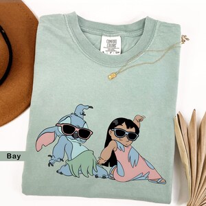 May include: A light green t-shirt with a graphic print of Stitch and Lilo from the Disney movie Lilo & Stitch. Stitch is wearing sunglasses and a green grass skirt. Lilo is wearing sunglasses and a pink dress with a blue and white pattern. The shirt has the text "COMFORT COLORS" printed on the tag.