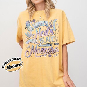 May include: Mustard-colored t-shirt with the words "Heels, Nails, Blade, Mascara" in a whimsical font. The design includes illustrations of a high heel, a hand with a butterfly, and a mascara tube. The words "Comfort Colors" and "Mustard" are also printed on the shirt.