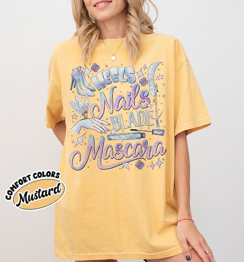 May include: Mustard-colored t-shirt with the words "Heels, Nails, Blade, Mascara" in a whimsical font. The design includes illustrations of a high heel, a hand with a butterfly, and a mascara tube. The words "Comfort Colors" and "Mustard" are also printed on the shirt.