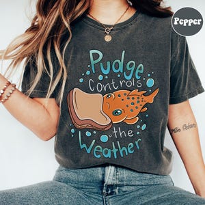 May include: A dark grey t-shirt with a cartoon illustration of a fish eating a peanut butter and jelly sandwich. The text "Pudge controls the weather" is printed on the shirt.