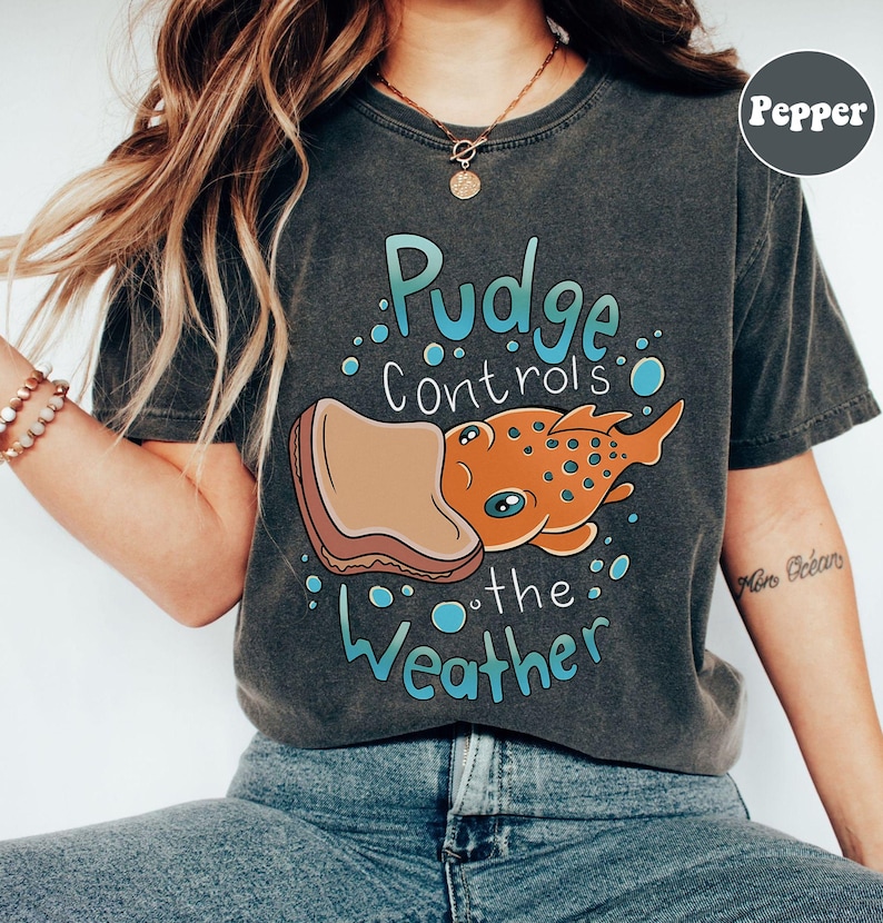 May include: A dark grey t-shirt with a cartoon illustration of a fish eating a peanut butter and jelly sandwich. The text "Pudge controls the weather" is printed on the shirt.