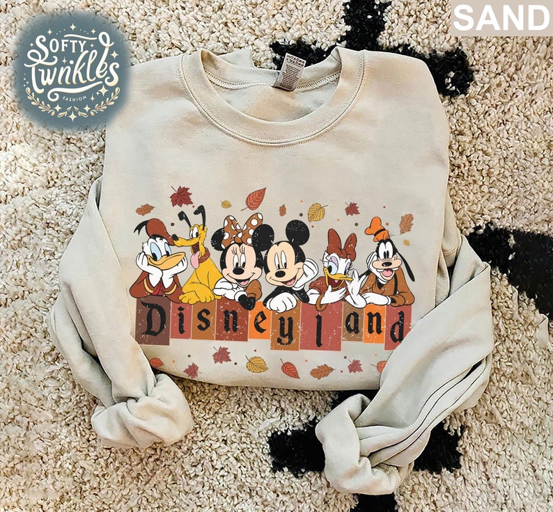 May include: A beige sweatshirt with a vintage-style graphic of Mickey Mouse, Minnie Mouse, Donald Duck, Goofy, and Pluto in front of a brick wall with the text "Disneyland" in brown, orange, and red. The characters are surrounded by autumn leaves.