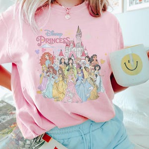 May include: Pink t-shirt with a graphic of Disney princesses in front of Cinderella's castle. The text "Disney Princess" is printed on the shirt.