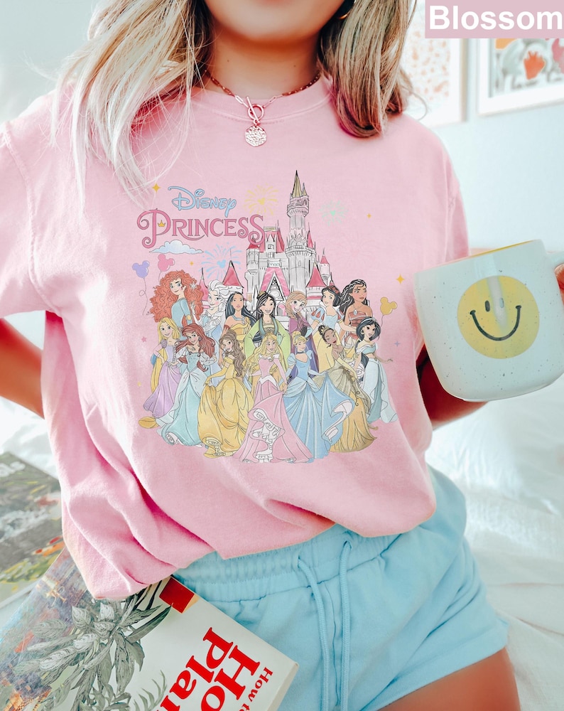 May include: Pink t-shirt with a graphic of Disney princesses in front of Cinderella's castle. The text "Disney Princess" is printed on the shirt.