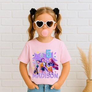 May include: A light pink t-shirt featuring a graphic of three animated characters, the number 5, and the name "MADISON" in glittery purple. The shirt is worn by a young girl with heart-shaped sunglasses and black hair ties.
