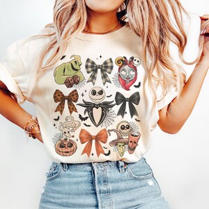 May include: A cream-coloured t-shirt with a design featuring Halloween-themed characters and bows. The design includes Jack Skellington, Sally, and Oogie Boogie, along with spiderwebs and bats.